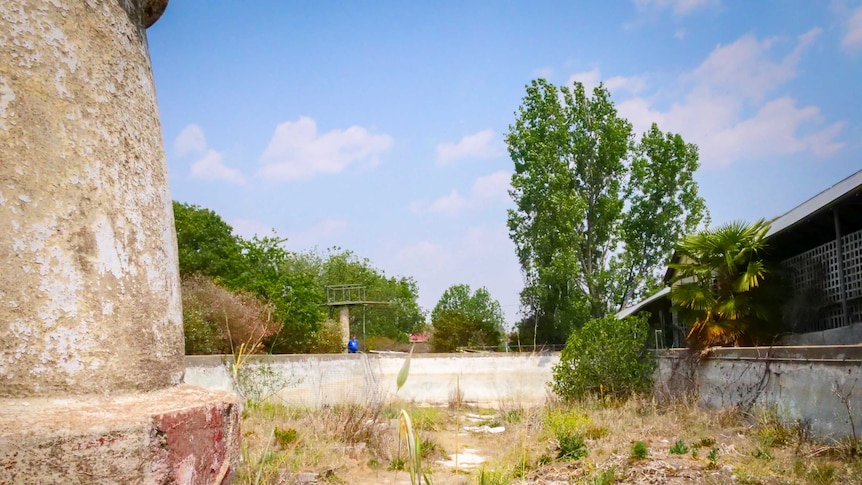 Looking the length of a derelict cement swimming pool, a stone fountain in the foreground and a diving board the other end