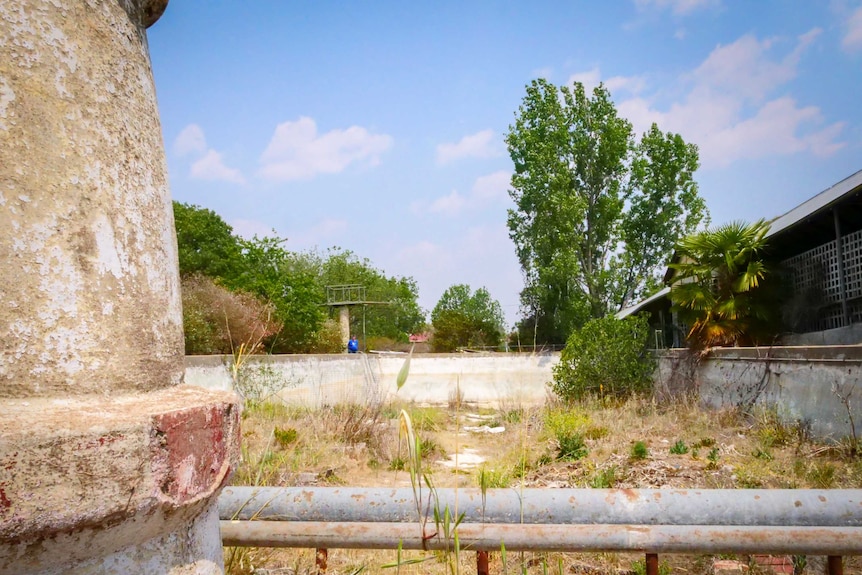 Looking the length of a derelict cement swimming pool, a stone fountain in the foreground and a diving board the other end