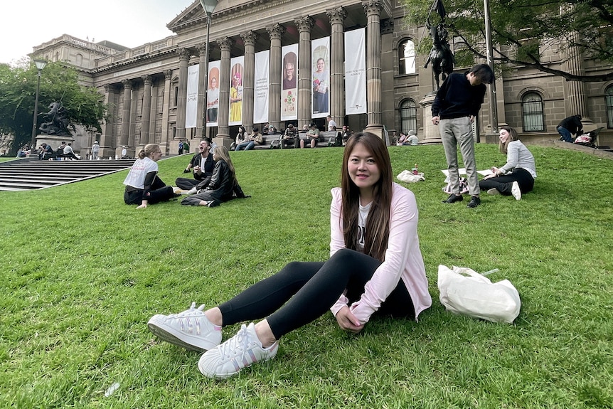 A smiling woman sits on a slanted lawn in front of a classical building.