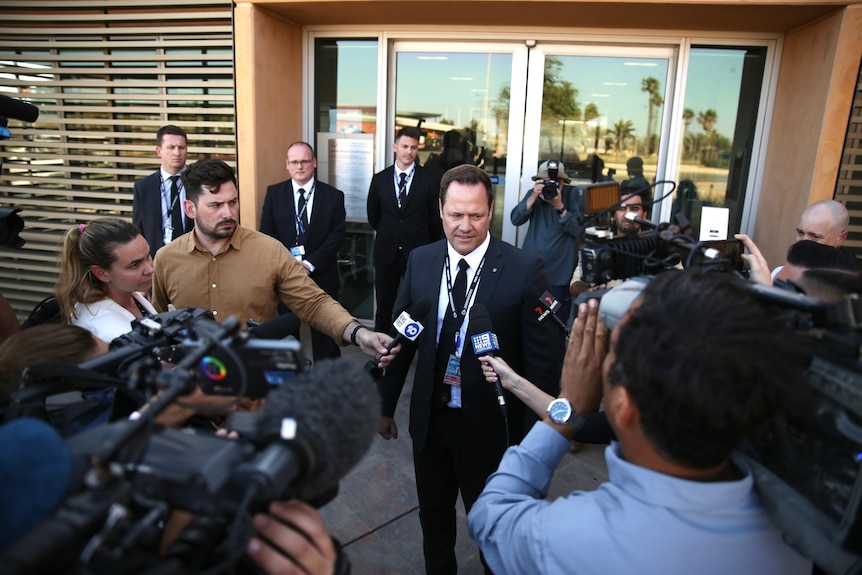 A detective in a black suit stands surrounded by dozens of reporters, camera operators, and photographers.