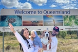 Alex standing in front of a welcome to QLD sign holding her daughter next to her husband who is holding their pet beagle