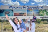 Alex standing in front of a welcome to QLD sign holding her daughter next to her husband who is holding their pet beagle