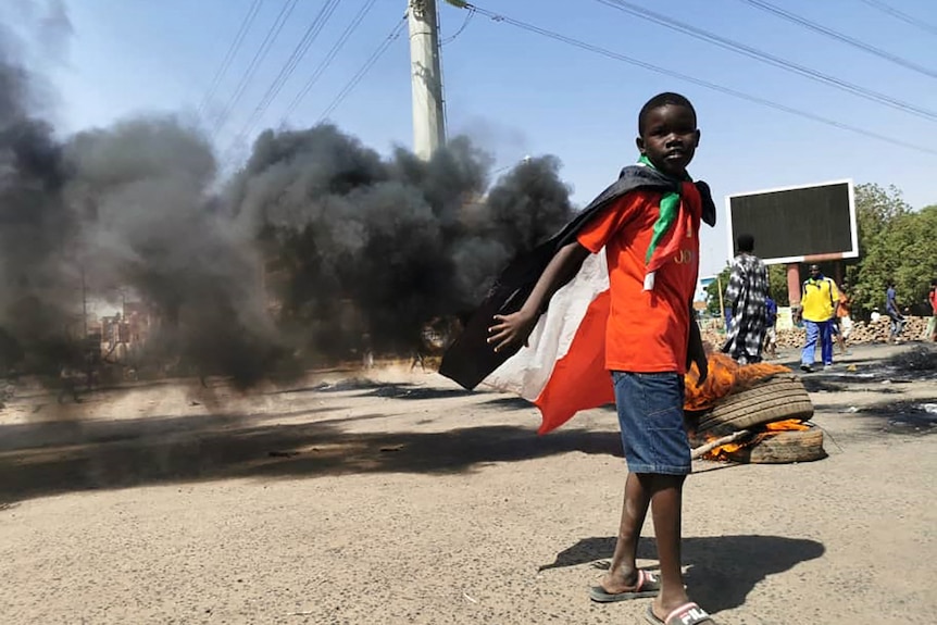 Un ragazzino nero vestito di arancione indossa la bandiera sudanese in una strada cittadina mentre il fumo si alza dietro di lui.