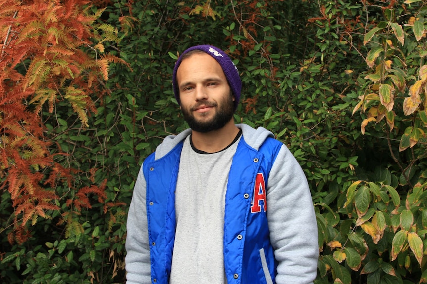 A young man wearing a beanie and bomber jacket stands in front of autumn coloured leaves.