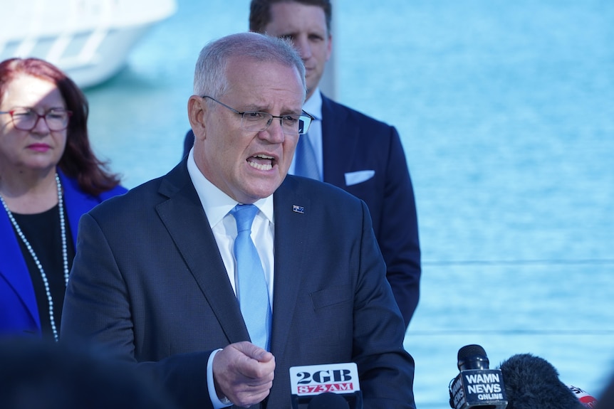 Scott Morrison, wearing a dark suit, gives a press conference outdoors at a shipyard, by the water.