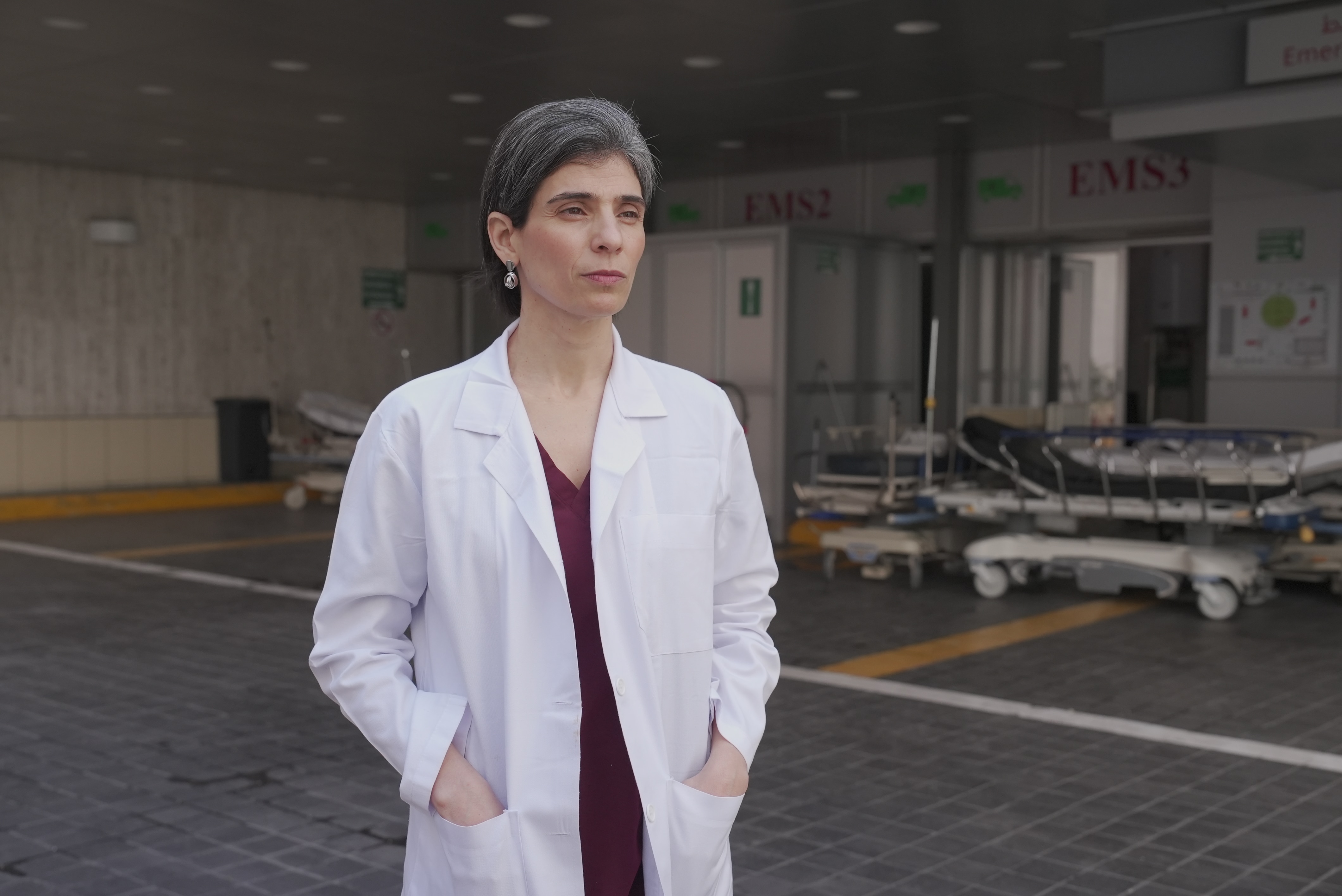 A doctor in a white coat looks solemn as she stands in front of a hospital.