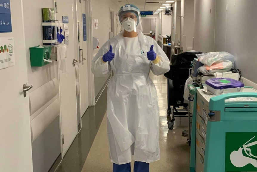 A woman in scrubs and personal protective gear gives the thumbs up signal.