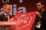 Mr Shorten is holding the banner the protester is brandishing, which says "Stop Adani"