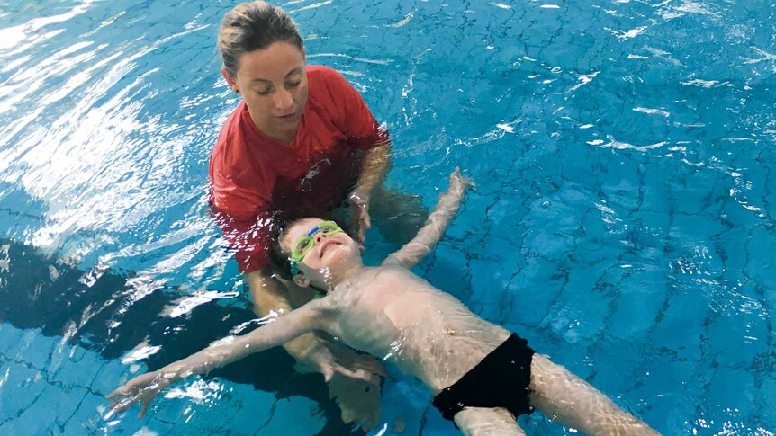 A woman in a red t-shirt holds a young boy as he floats face up in a pool, arms outstretched.