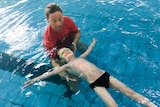 A woman in a red t-shirt holds a young boy as he floats face up in a pool, arms outstretched.