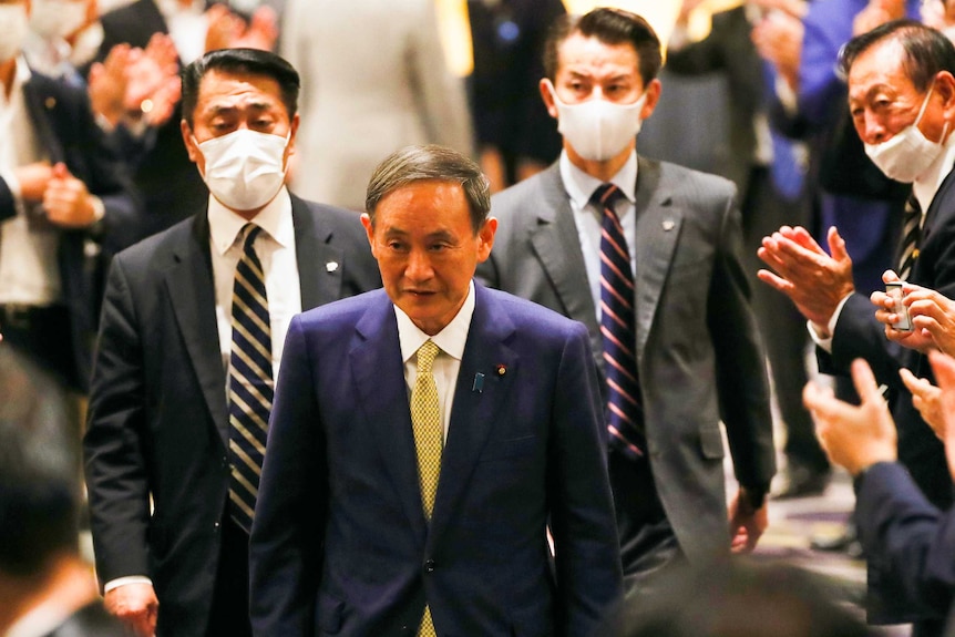 Yoshihide Suga walking in a room filled with men wearing face masks