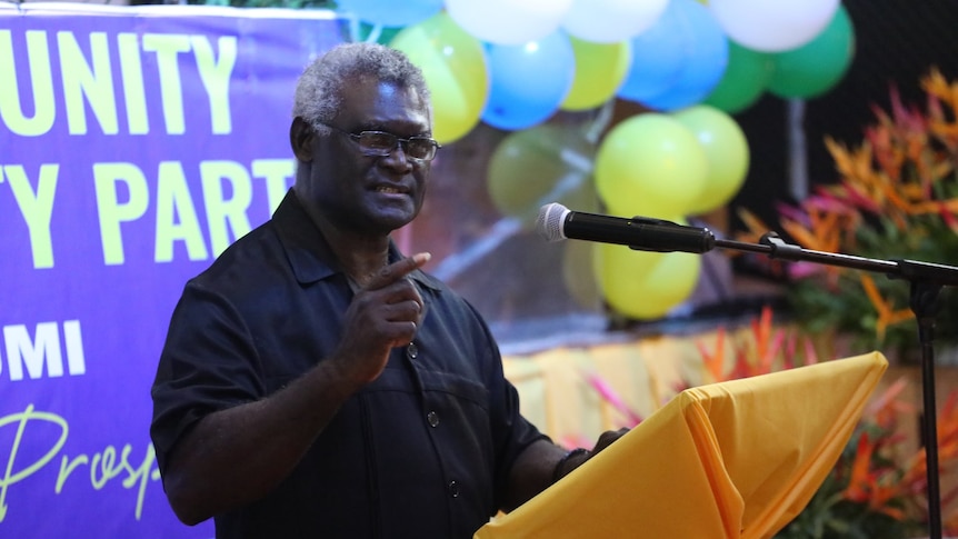 A man gestures as he speaks on a stage in front of balloons and a political banner