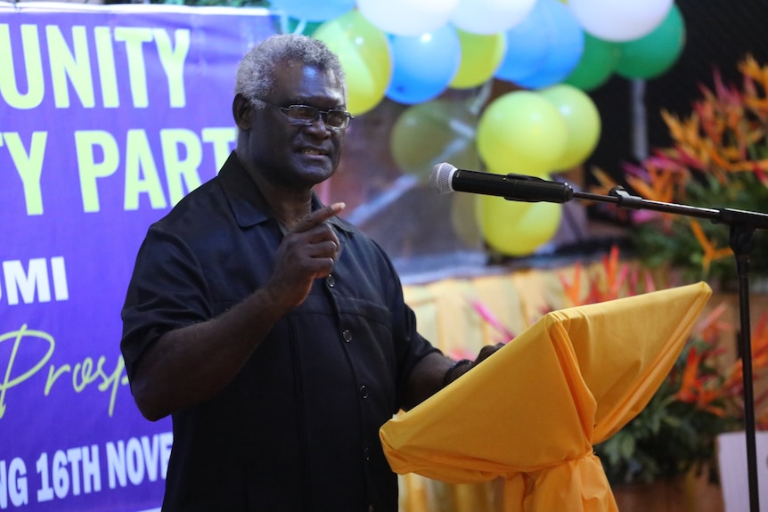 A man gestures as he speaks on a stage in front of balloons and a political banner