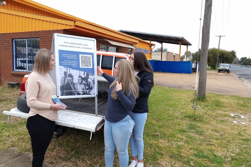 Three women stand around the tray of a ute carrying a sign with a picture of a man and woman on it.