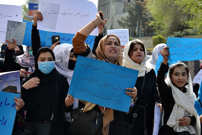 A group of Muslim women in hijabs and face masks hold blue signs with Arabic writing in outdoor protest