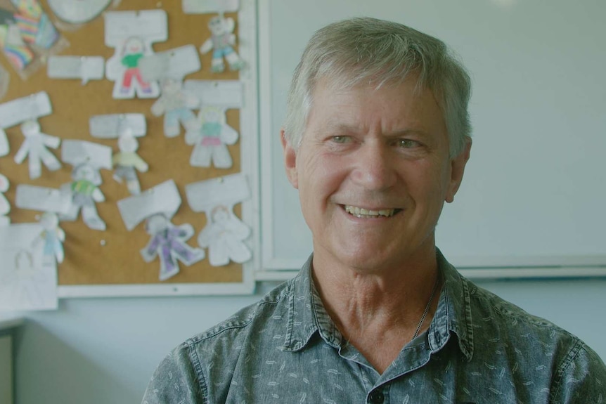 Man smiling in classroom in front of whiteboard, childrens' artwork in background