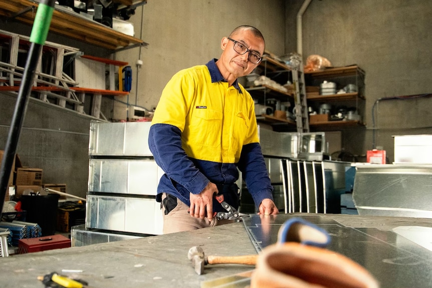 Yaning Tang wearing a high-vis shirt in a workshop, cutting sheets of steel and looking toward the camera.