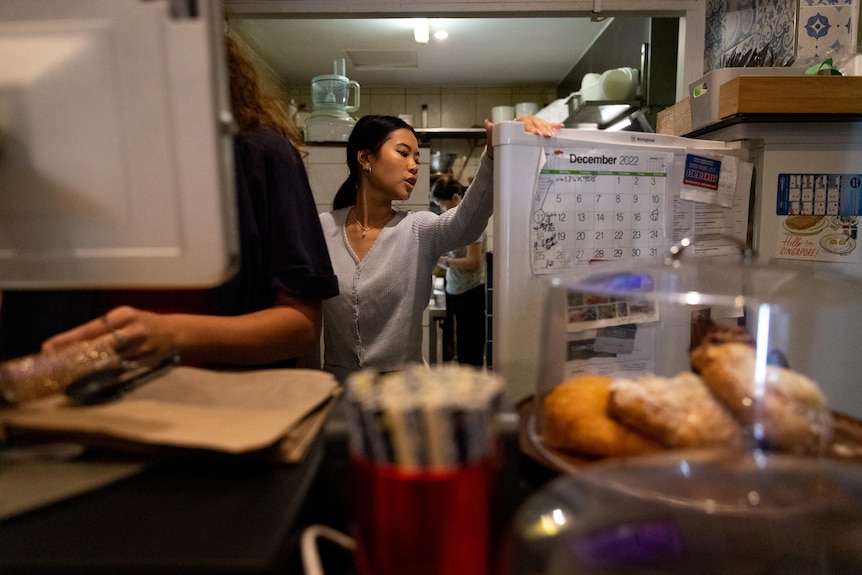 A teenage girl opens a fridge behind a cluttered counter in a small cafe.
