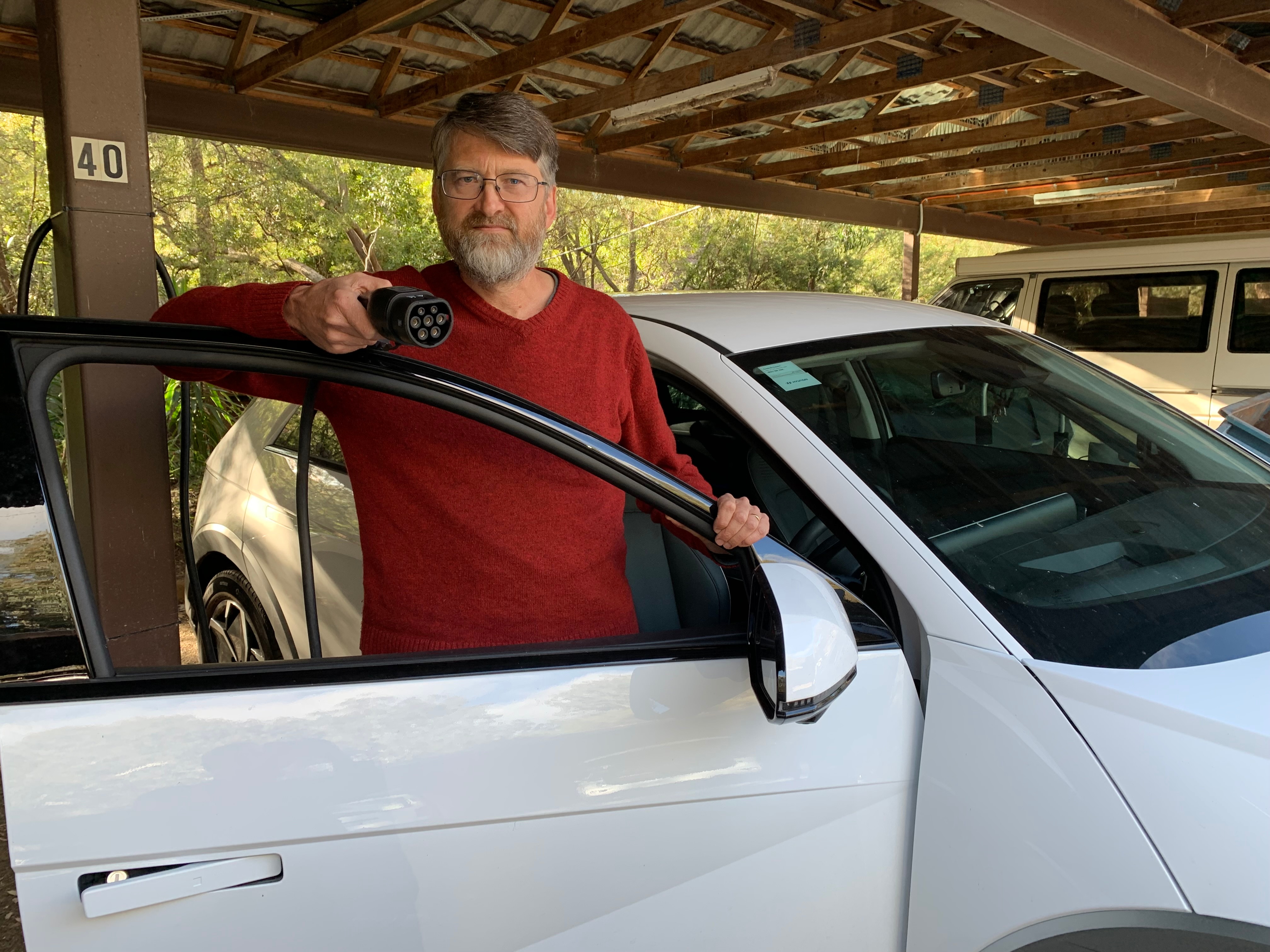 A man stands next to his electric vehicle pointing the car charger toward the camera. 
