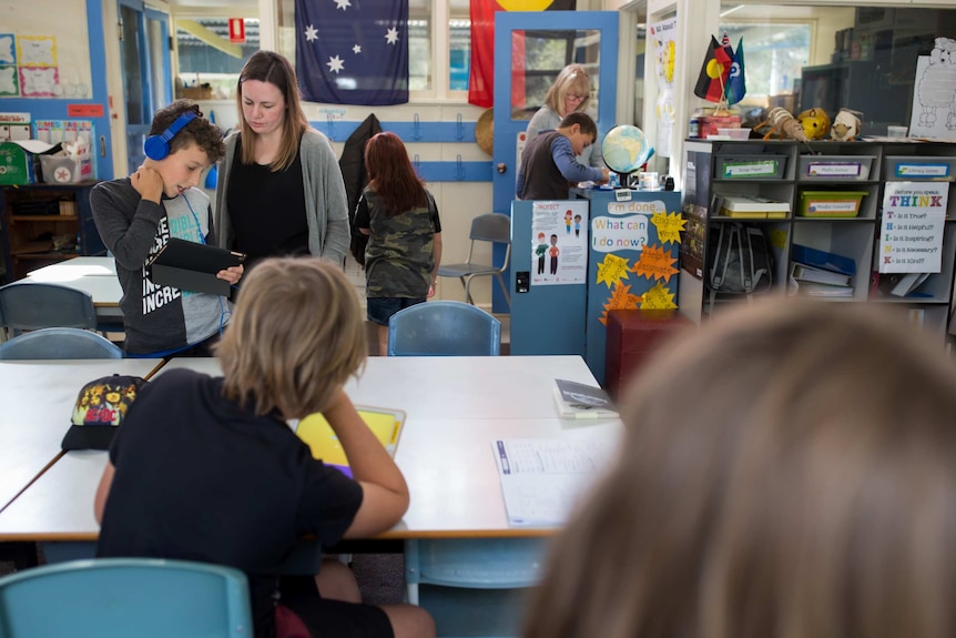 Teacher Anita Harding helps a student working with headphones on an iPad, surrounded by classmates in a colourful classroom.