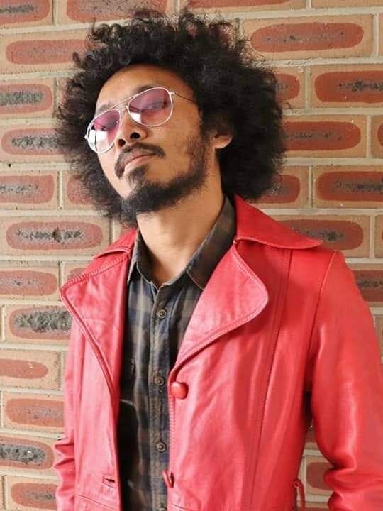 A man with curly big hair, beard and moustache, wearing brown jacket and red leather jacket and glasses, posing to the camera.