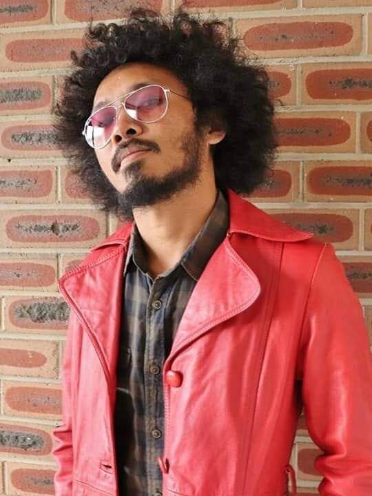 A man  with curly big hair, beard and moustache, wearing brown jacket and red leather jacket and glasses, posing to the camera.