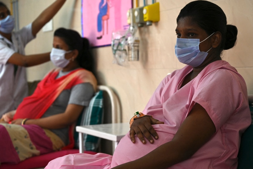 A pregnant woman wearing pink surgical clothes and a wask waits on a hospital bed. 