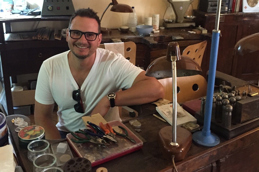 A man with glasses smiles as he sits at a desk with tools.