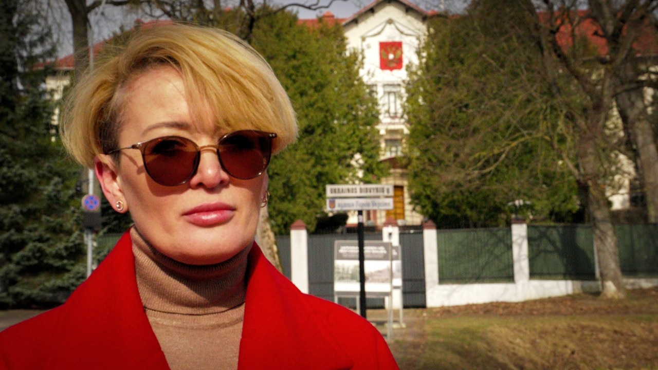 A woman with sunglases on looks at the camera, outside a large building with a fence around it