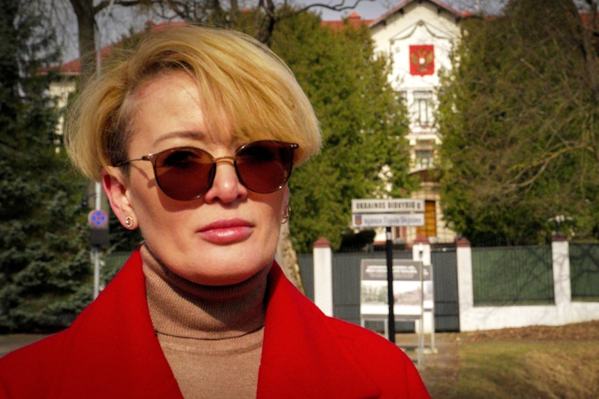 A woman with sunglases on looks at the camera, outside a large building with a fence around it