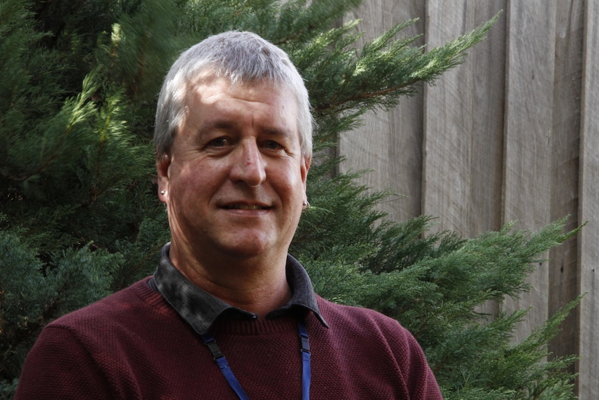 a portrait shot of a man wearing a burgundy jumper and dark collared shirt in a garden