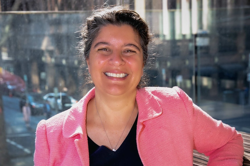 Woman in pink jacket looks at camera, smiling, city street in background