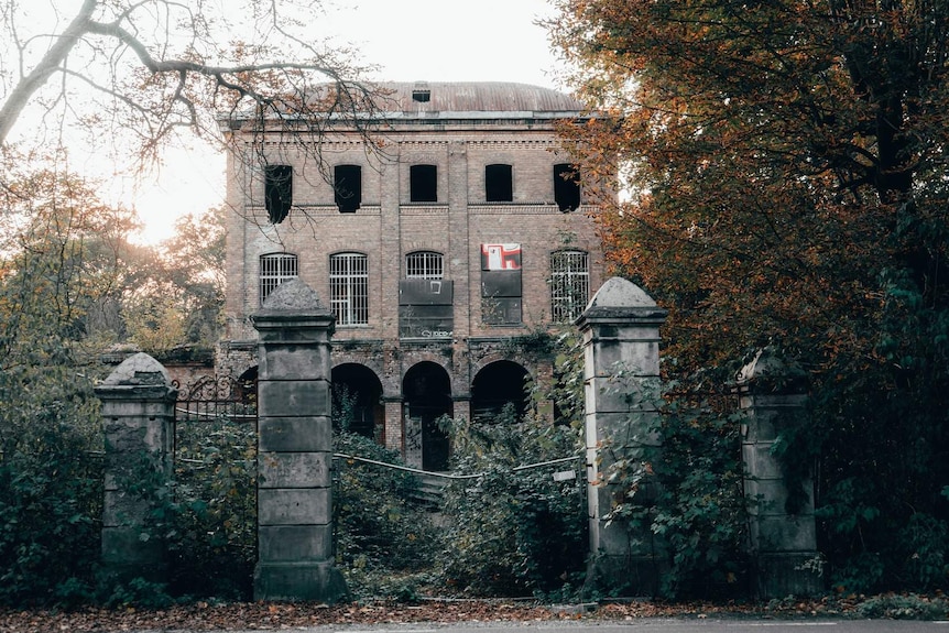 An old, dilapidated brick building in Germany with a fallen down fence and overgrown vegetation