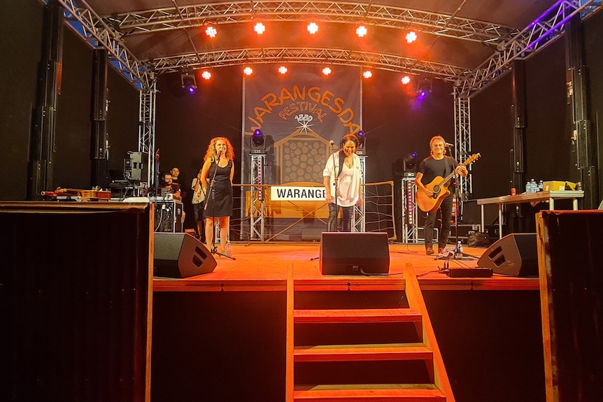 Three singers lined up on a stage with orange and red lights behind them.