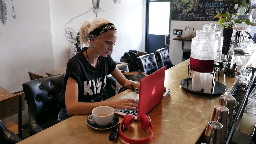 Blonde woman with KISS t-shirt sits at cafe counter with red laptop open.