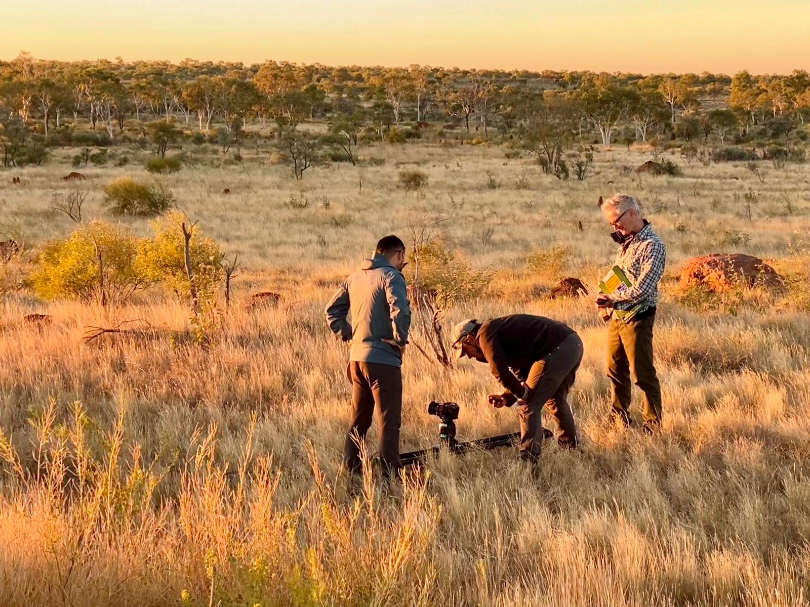 Three men looking at camera and sound equipment in bush landscape.