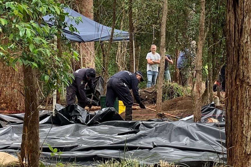 Two police officers digging and raking in a cleared patch of woodland while several people look on.  