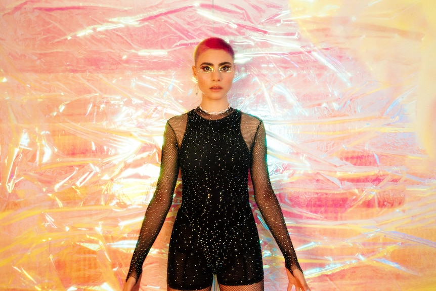 A woman stands in front of an iridescent background while wearing a black, sparkly jumpsuit. Her hair is dyed.