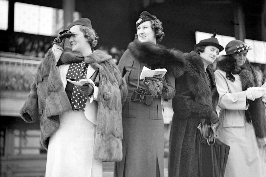 Women in fur stand in front of a grandstand, one looks through binoculars.