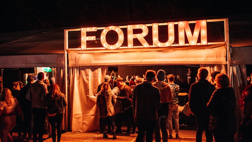 A crowd of people stand outside Splendour's Forum tent under the lit-up sign at night.