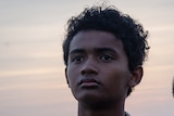 A boy wearing white patterned button-up shirt looks in the distance with solemn expression in front of a dusk or sunset sky.