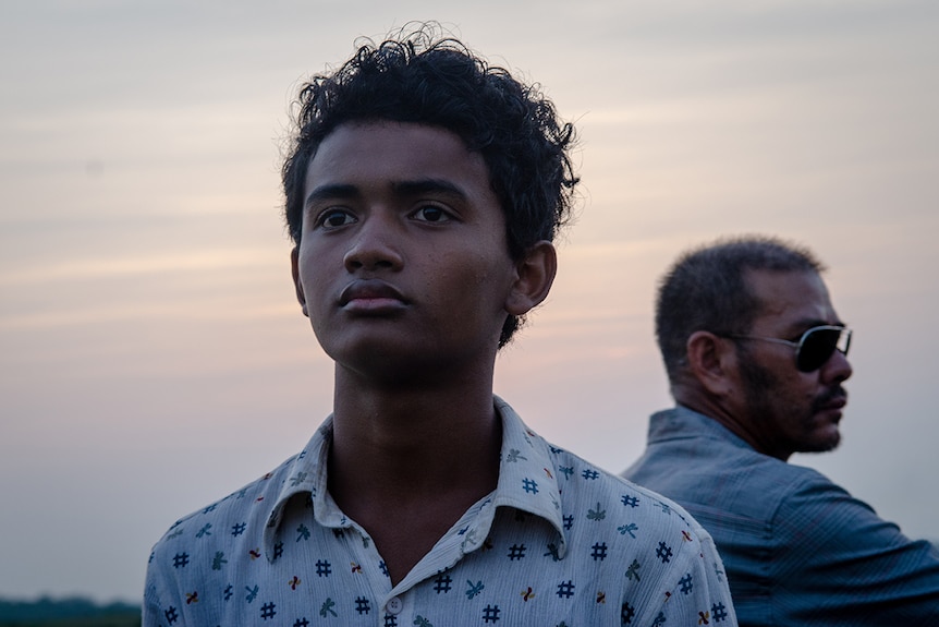 A boy wearing white patterned button-up shirt looks in the distance with solemn expression in front of a dusk or sunset sky.