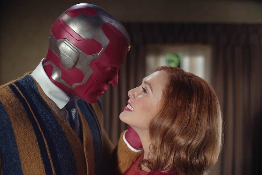 A 30-something woman looks up at a masked man. She is smiling lovingly and craning her neck up. Both wear 60s-style clothing.