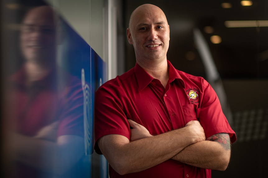 A bald man smiles at the camera with his arms crossed. He's wearing a red collared T-shirt.