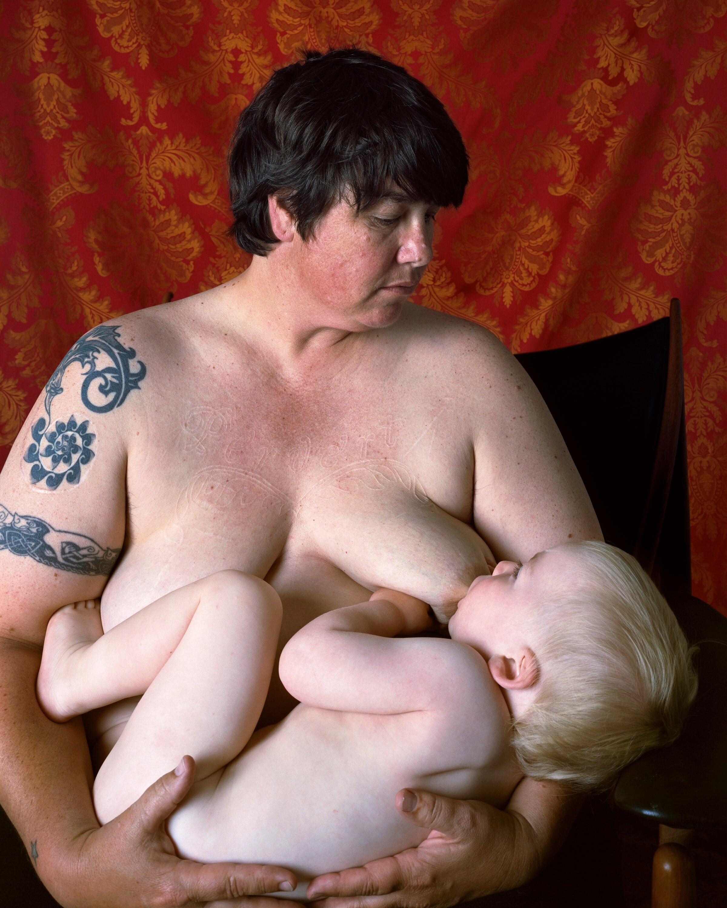 A bare-chested woman, with short brown hair and tattoos on her right shoulder, gazes at her baby as she nurses him