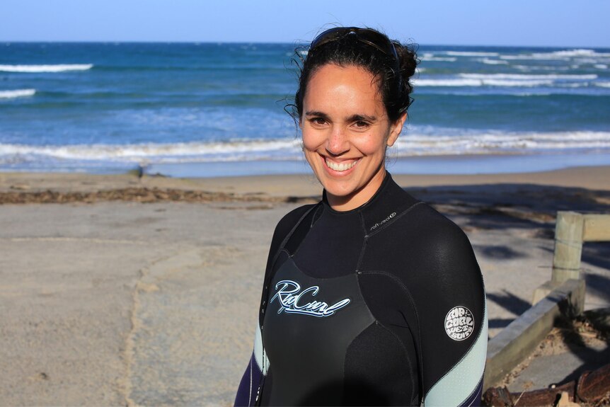 A woman with dark brown curly hair smiles at the camera as she stands on the beach in a Ripcurl wetsuit.