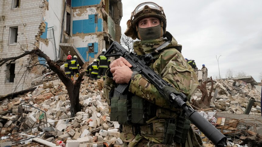 A soldier in military uniform and holding a gun stands in front of a destroyed apartment building with emergency crews.