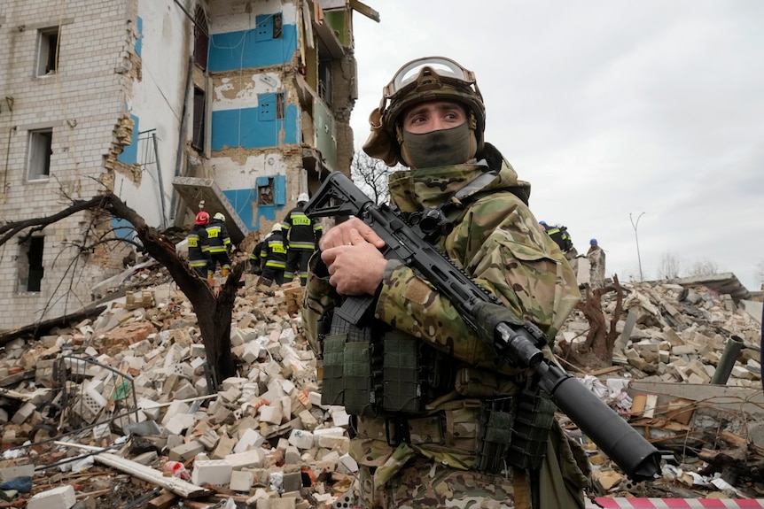 A soldier in military uniform and holding a gun stands in front of a destroyed apartment building with emergency crews.