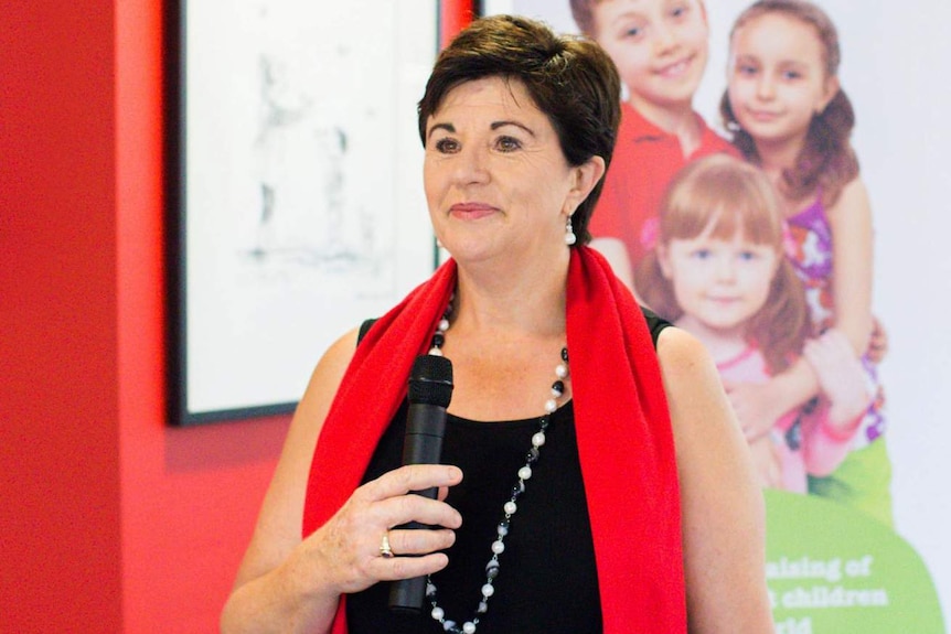 A mid shot of Maggie Dent speaking in a room holding a microphone and wearing a black top and red scarf.