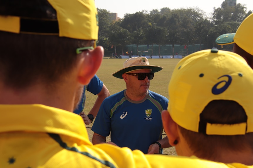 Jason Mathers stands and talks to men wearing yellow cricket kit.